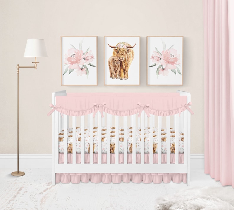 May include: A nursery set featuring a white crib with pink bedding, including a ruffled bumper and skirt. Above the crib are three framed prints: two with pink floral designs and one with a watercolor illustration of a Highland cow and calf.