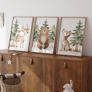 Op de afbeelding: Drie ingelijste aquarelprints met bosdieren: een hert, een beer en een vos. Elke print heeft een licht houten frame en toont de dieren met dennenbomen op de achtergrond. De prints worden weergegeven op een donkere houten kast.
