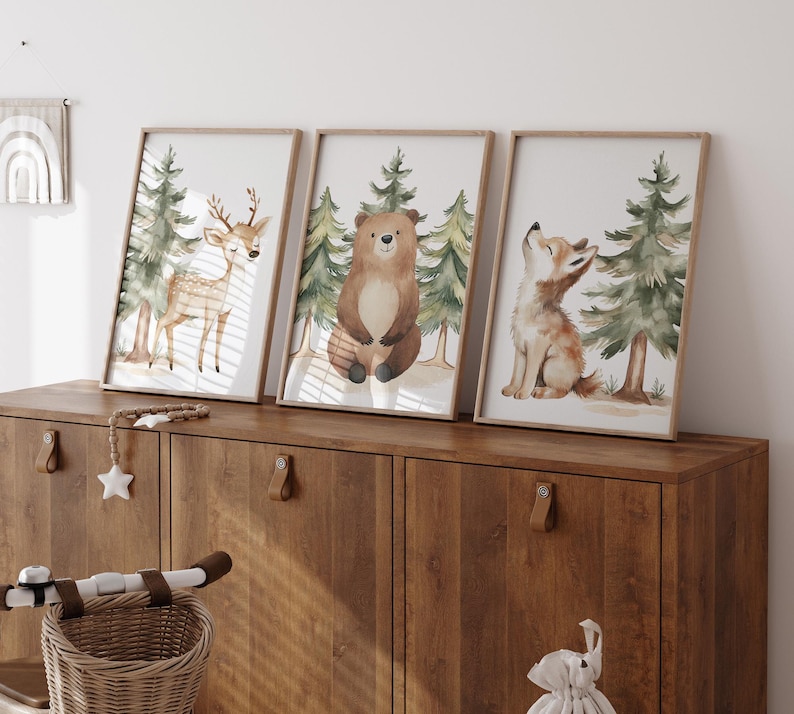 Op de afbeelding: Drie ingelijste aquarelprints met bosdieren: een hert, een beer en een vos. Elke print heeft een licht houten frame en toont de dieren met dennenbomen op de achtergrond. De prints worden weergegeven op een donkere houten kast.