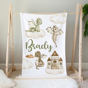 Puede incluir: Una manta de bebé blanca con un diseño de dragón verde y marrón. La manta está personalizada con el nombre "Brady" e incluye una ilustración de un castillo.