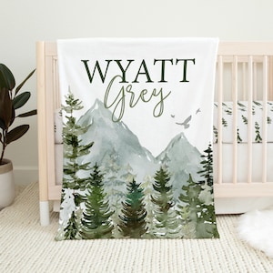 Puede incluir: Una manta de bebé blanca con una ilustración de acuarela de una cordillera y pinos. El texto "WYATT Grey" está impreso en verde en la parte superior de la manta.