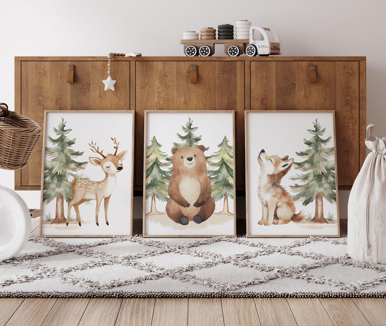Op de afbeelding: Drie ingelijste aquarelprints met bosdieren: een hert, een beer en een wolf. Elke print heeft een lichte achtergrond met groene bomen. De prints worden tentoongesteld op een houten kast boven een grijs tapijt.