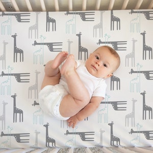 May include: A baby lying on a white crib sheet with a gray and light blue giraffe print. The baby is wearing a white onesie.