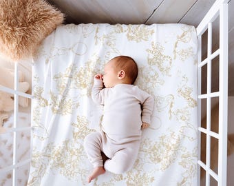 May include: A sleeping baby in a white cot. The baby wears a light beige onesie and lies on a white sheet with a gold animal print. A fluffy beige pillow is in the upper left corner of the cot.