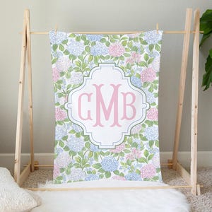 Puede incluir: Una manta rectangular con un patr&oacute;n repetitivo de flores de hortensia azules y rosas con hojas verdes. En el centro, un marco blanco en forma de cuatrifolio contiene las grandes letras de monograma GMB en color rosa y estilo serif.