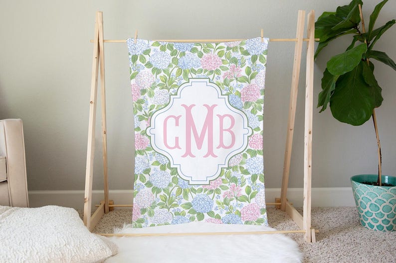 Puede incluir: Una manta rectangular con un patr&oacute;n repetitivo de flores de hortensia azules y rosas con hojas verdes. En el centro, un marco blanco en forma de cuatrifolio contiene las grandes letras de monograma GMB en color rosa y estilo serif.