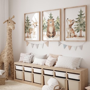 Op de afbeelding: Een kinderkamer met drie ingelijste aquarelprints van bosdieren: een hert, een beer en een vos. Daaronder staat een houten opbergmeubel met witte bakken, versierd met kussens. Een grote geweven giraf staat links.