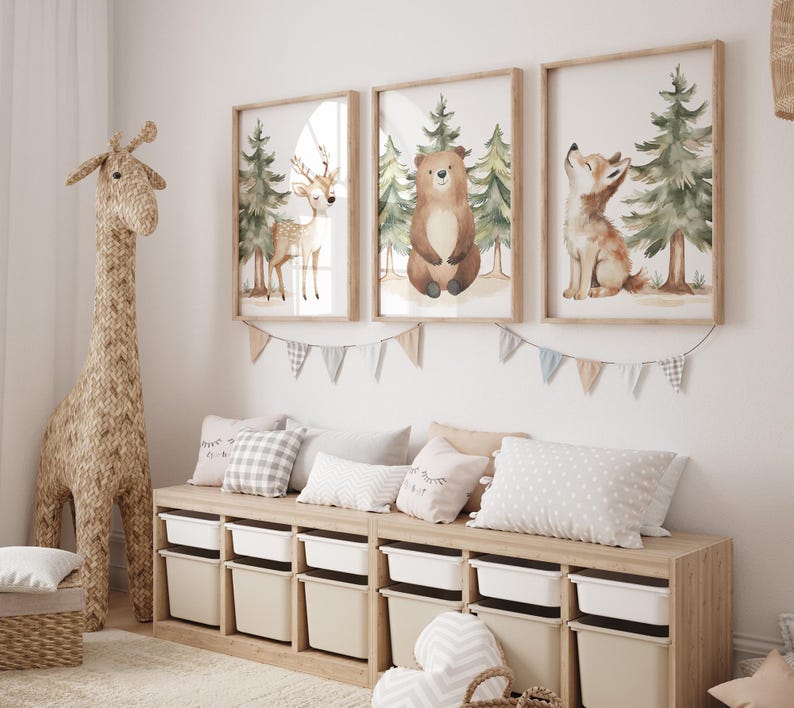 Op de afbeelding: Een kinderkamer met drie ingelijste aquarelprints van bosdieren: een hert, een beer en een vos. Daaronder staat een houten opbergmeubel met witte bakken, versierd met kussens. Een grote geweven giraf staat links.