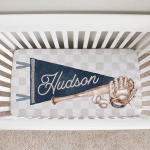 Puede incluir: Una sábana de cuna con un patrón de cuadros gris claro y blanco. En el centro, un banderín azul marino muestra el nombre Hudson en letra cursiva blanca, junto a un bate de béisbol de madera, un guante de cuero y dos pelotas de béisbol.