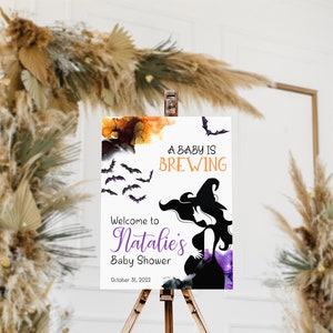Op de afbeelding: Een wit bord met een paarse en oranje aquarel achtergrond. Het bord zegt "A baby is brewing" met vleermuizen die rond de tekst vliegen. Onder de tekst is een silhouet van een zwangere vrouw die een heksenhoed draagt. Het bord zegt ook "Welcome to Natalie's Baby Shower" en "October 31, 2022".
