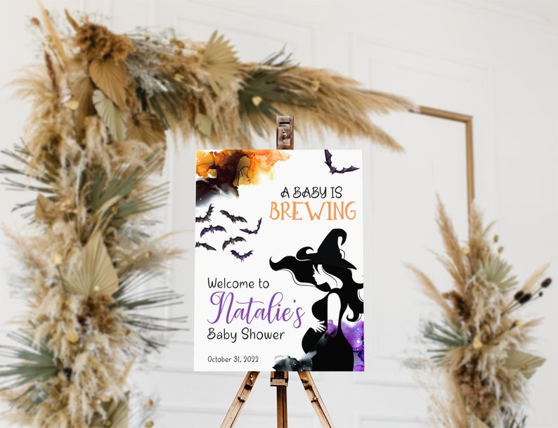 Op de afbeelding: Een wit bord met een paarse en oranje aquarel achtergrond. Het bord zegt "A baby is brewing" met vleermuizen die rond de tekst vliegen. Onder de tekst is een silhouet van een zwangere vrouw die een heksenhoed draagt. Het bord zegt ook "Welcome to Natalie's Baby Shower" en "October 31, 2022".