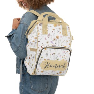 May include: A white canvas backpack with a floral print and a beige top flap. The backpack has a large zippered pocket on the front with a beige label that says "Hannah".