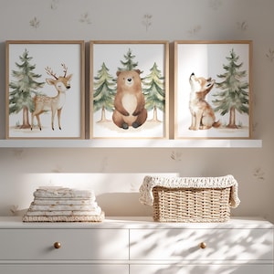 Op de afbeelding: Drie ingelijste aquarelprints met bosdieren: een hert, een beer en een wolf. Elke print heeft een licht houten frame en toont de dieren met dennenbomen op de achtergrond. De prints worden weergegeven boven een witte dressoir met opgevouwen linnen en een rieten mand.