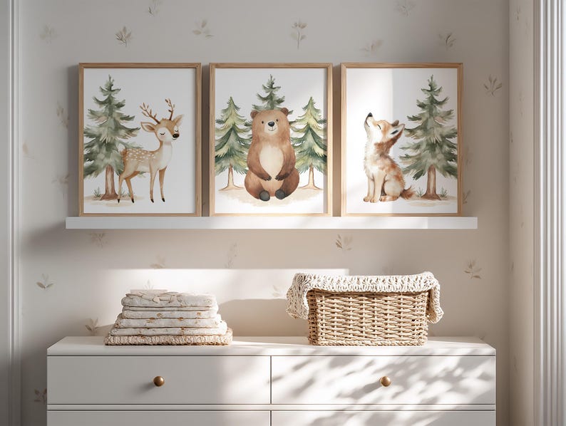 Op de afbeelding: Drie ingelijste aquarelprints met bosdieren: een hert, een beer en een wolf. Elke print heeft een licht houten frame en toont de dieren met dennenbomen op de achtergrond. De prints worden weergegeven boven een witte dressoir met opgevouwen linnen en een rieten mand.