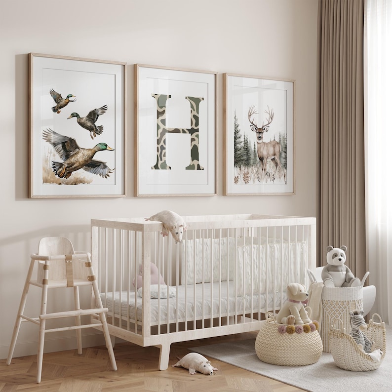May include: Three framed art prints with nature themes hang above a white crib. The prints feature ducks in flight, a camouflage-patterned letter "H", and a deer in a forest. A high chair and baskets with stuffed animals complete the nursery setting.
