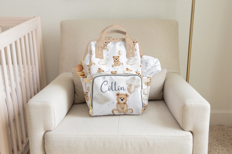 K&ouml;nnte beinhalten: Eine wei&szlig;e Wickeltasche mit einem braunen Teddyb&auml;r-Druck und dem Namen "Colton" bestickt auf der Vorderseite. Die Tasche hat einen beigefarbenen Griff und steht auf einem hellbeigen Sessel.