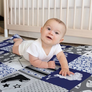 May include: A baby is lying on a blue and gray patchwork baby blanket with hockey-themed designs. The blanket features a variety of patterns, including stars, stripes, and hockey sticks. The text "GOAL" is visible on the blanket.