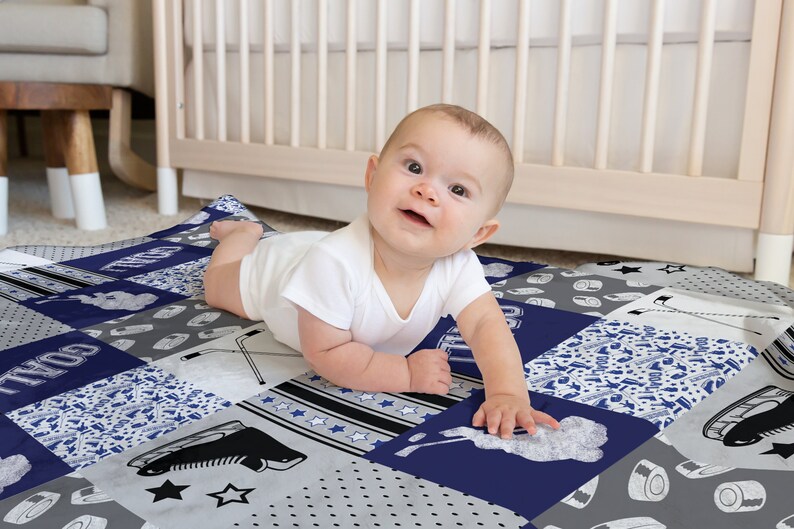 May include: A baby is lying on a blue and gray patchwork baby blanket with hockey-themed designs. The blanket features a variety of patterns, including stars, stripes, and hockey sticks. The text "GOAL" is visible on the blanket.