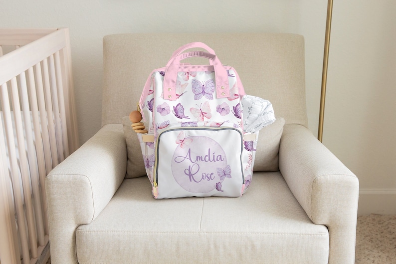May include: A white nappy bag with a pink butterfly pattern and the name "Amelia Rose" printed on the front. The bag has a pink zipper and a pink handle.