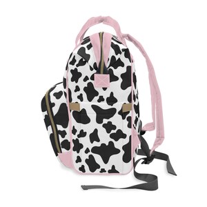 May include: A black and white cow print backpack with pink accents. The backpack has a large main compartment and a smaller front pocket. The straps are adjustable and the backpack is made of a durable fabric.