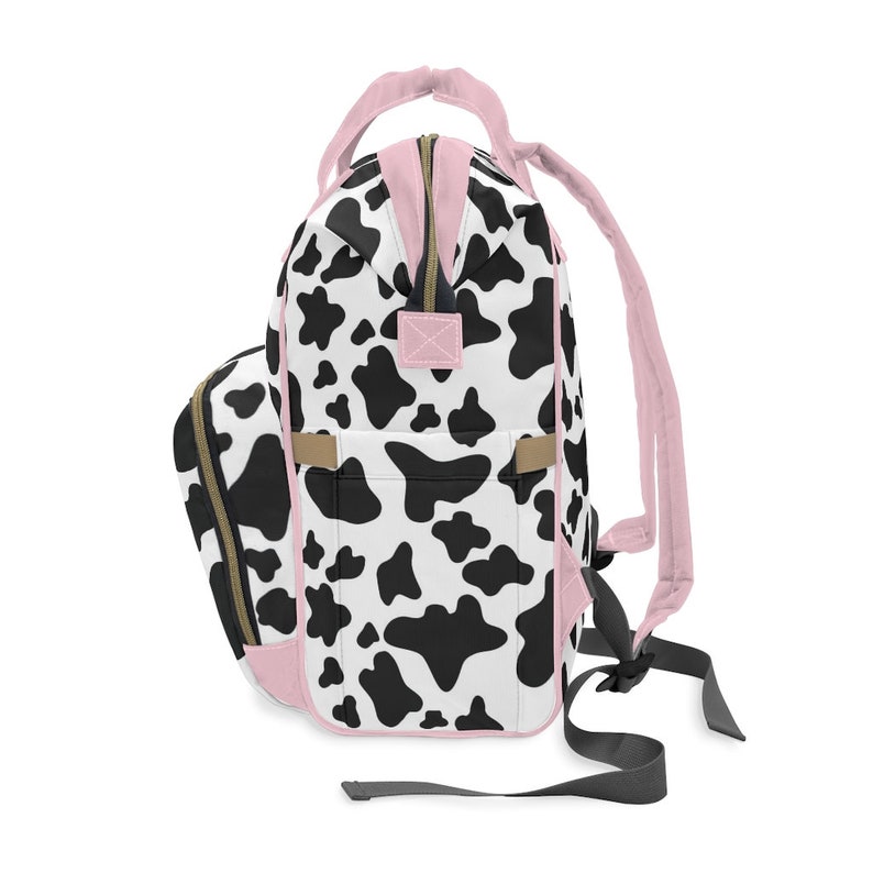 May include: A black and white cow print backpack with pink accents. The backpack has a large main compartment and a smaller front pocket. The straps are adjustable and the backpack is made of a durable fabric.