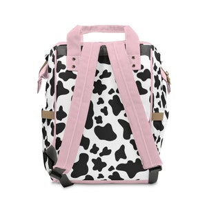 May include: A pink and black cow print backpack with adjustable straps. The backpack has a top handle and a zipped pocket on the front.