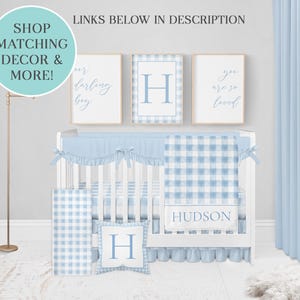 May include: A white crib with a blue and white gingham crib bedding set. The bedding includes a fitted sheet, a blanket, a pillow, and a crib skirt. The crib is decorated with three framed prints, one with the letter "H" and two with text. The text on the prints reads "Our darling boy" and "You are so loved".