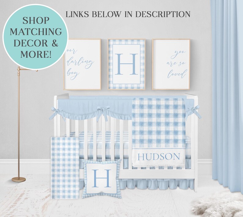 May include: A white crib with a blue and white gingham crib bedding set. The bedding includes a fitted sheet, a blanket, a pillow, and a crib skirt. The crib is decorated with three framed prints, one with the letter "H" and two with text. The text on the prints reads "Our darling boy" and "You are so loved".