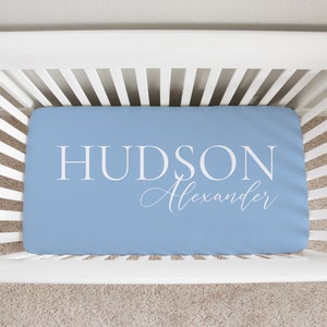 以下が含まれることがあります： 大きな白い大文字で「HUDSON」、筆記体で「Alexander」と書かれた、水色のベビーベッドシーツ。白いベビーベッドの中にシーツがあります。