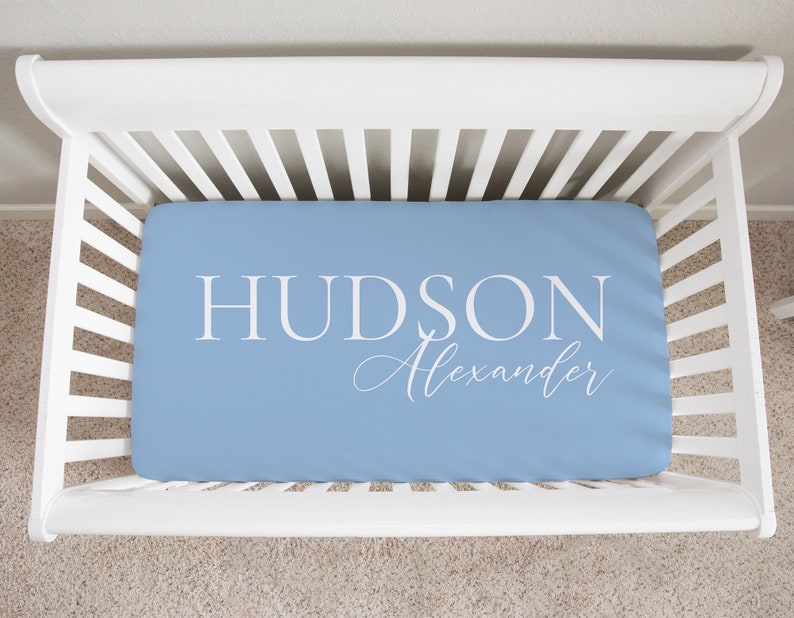 以下が含まれることがあります： 大きな白い大文字で「HUDSON」、筆記体で「Alexander」と書かれた、水色のベビーベッドシーツ。白いベビーベッドの中にシーツがあります。