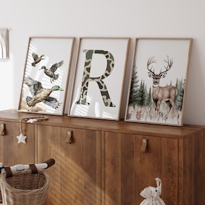 May include: Three framed art prints featuring wildlife. One print shows ducks in flight, another displays a camouflage-patterned letter "R", and the third depicts a deer in a forest setting. The prints are displayed on a wooden cabinet.