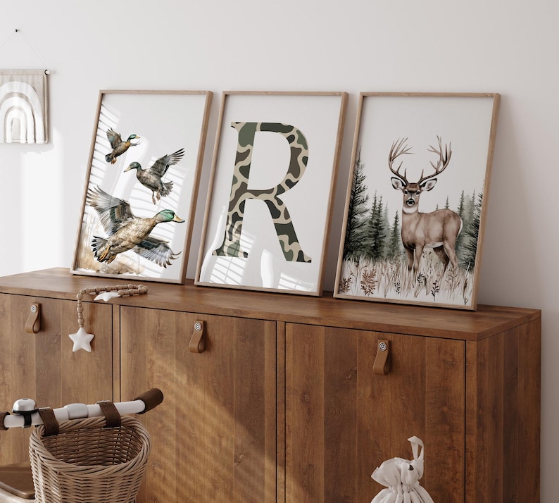 May include: Three framed art prints featuring wildlife. One print shows ducks in flight, another displays a camouflage-patterned letter "R", and the third depicts a deer in a forest setting. The prints are displayed on a wooden cabinet.
