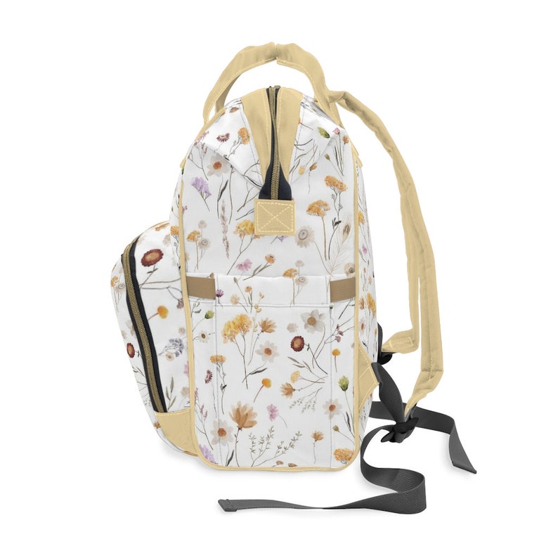 May include: A white backpack with a floral pattern featuring dried flowers in shades of yellow, orange, and pink. The backpack has a cream-colored zipper and straps.