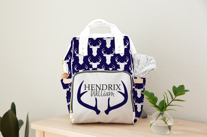 May include: A white diaper bag with a navy blue deer print and gold accents. The bag has a large front pocket with the names "Hendrix William" and a pair of antlers printed on it.