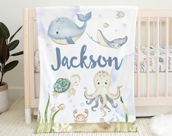 May include: A white baby blanket with a blue whale, octopus, turtle, and stingray. The blanket has a light blue background with a light blue and white watercolor design. The blanket also has the name "Jackson" written in blue letters.