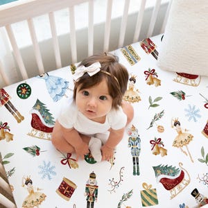 Könnte beinhalten: Ein Baby in einem weißen Body sitzt auf einem Bettlaken mit festlichem Weihnachts-Nussknacker-Motiv. Das Laken zeigt Illustrationen von Nussknackern, Ballerinas, Schlitten und Ornamenten in Rot, Grün und Gold auf weißem Hintergrund.