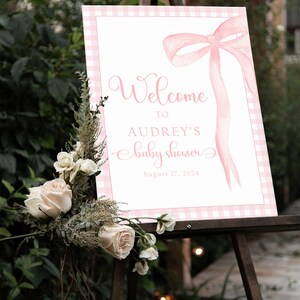 May include: A pink and white gingham welcome sign with a pink watercolor bow and the text "Welcome to Audrey's baby shower August 17, 2024".