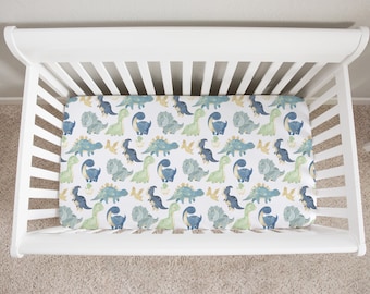 May include: White crib with a fitted sheet featuring a dinosaur pattern. The sheet has a white background with blue, green, and yellow dinosaurs.
