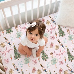Könnte beinhalten: Ein Baby liegt auf einem rosafarbenen Bettlaken mit einem sich wiederholenden Muster aus Weihnachtsbäumen, Ballerinas, Nussknackern, Schneeflocken und Schleifen. Der Name "Sophia" ist kursiv geschrieben. Das Baby trägt einen weißen Body und eine weiße Schleife.