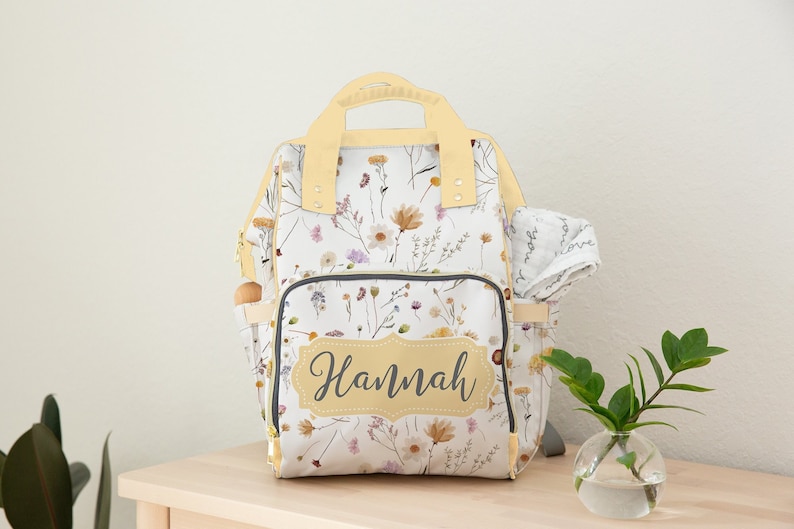 May include: A white diaper bag with a floral print and a yellow handle. The bag has a large pocket on the front with the name "Hannah" written in a script font.