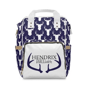 May include: White diaper bag backpack with a navy blue deer print. The front of the bag has a white deer antler design with the text "HENDRIX William" in black and gray.