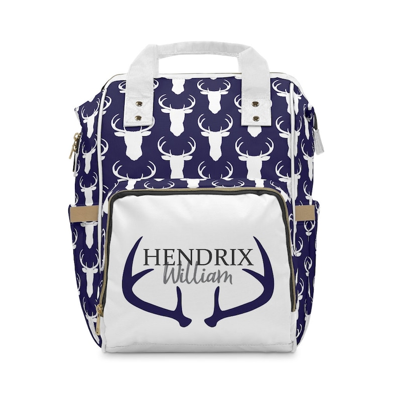 May include: White diaper bag backpack with a navy blue deer print. The front of the bag has a white deer antler design with the text "HENDRIX William" in black and gray.