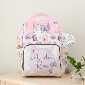 May include: A white nappy bag with a pink handle and a purple butterfly pattern. The bag has a front pocket with a purple circle and the text "Amelia Rose" in purple script.