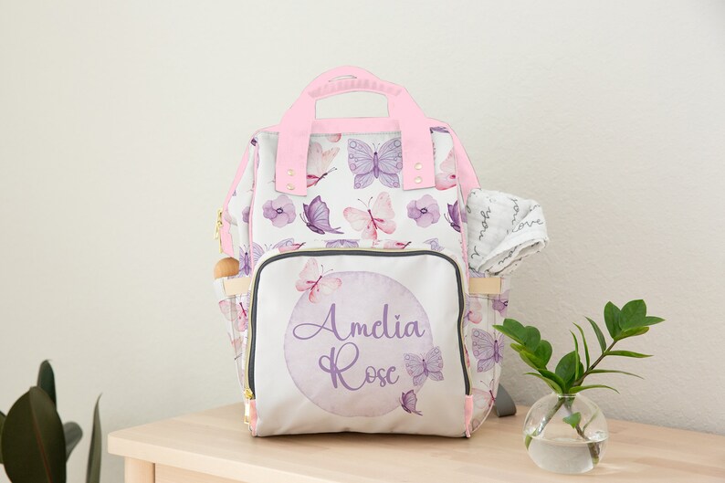 May include: A white nappy bag with a pink handle and a purple butterfly pattern. The bag has a front pocket with a purple circle and the text "Amelia Rose" in purple script.