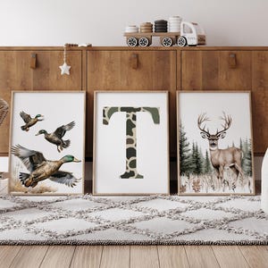 May include: Three framed art prints. One features flying ducks, another a camouflage letter "T", and the third a deer in a forest. The prints are displayed on a wooden cabinet with a toy train and decorative star.