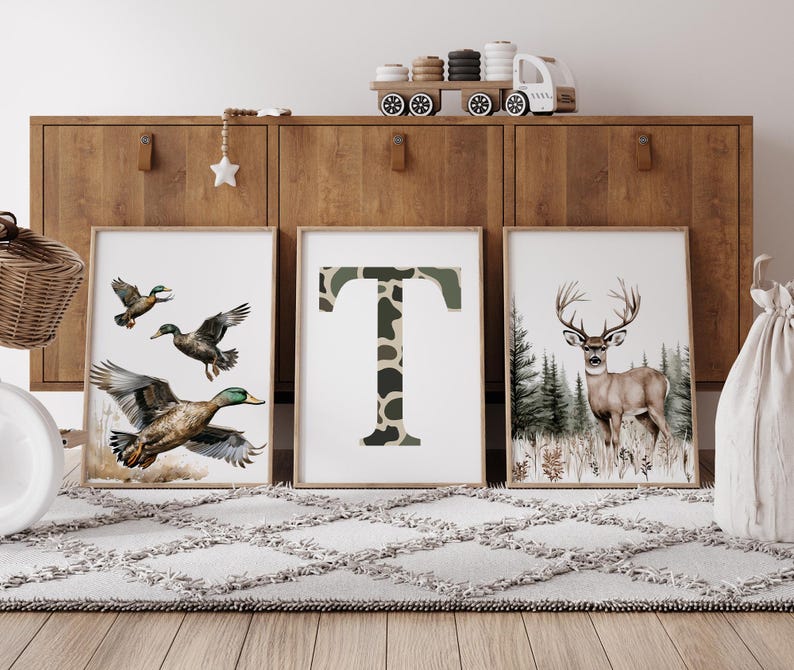 May include: Three framed art prints. One features flying ducks, another a camouflage letter "T", and the third a deer in a forest. The prints are displayed on a wooden cabinet with a toy train and decorative star.