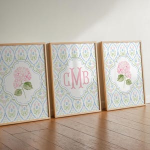 May include: Three rectangular framed wall art prints with light wood frames. The prints feature a repeating blue and pink floral damask pattern. The center print displays a pink monogram with the letters CMB, while the side prints show pink hydrangea flowers.
