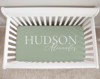 May include: A sage green fitted cot sheet with white text that reads "HUDSON Alexander".