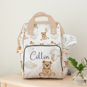 K&ouml;nnte beinhalten: Eine wei&szlig;e Wickeltasche mit einem braunen Teddyb&auml;r- und Ballon-Druck. Die Tasche hat eine Rei&szlig;verschlusstasche mit dem Namen "Colton" darauf. Die Tasche steht auf einem Holztisch mit einer kleinen Pflanze in einer Glasvase.