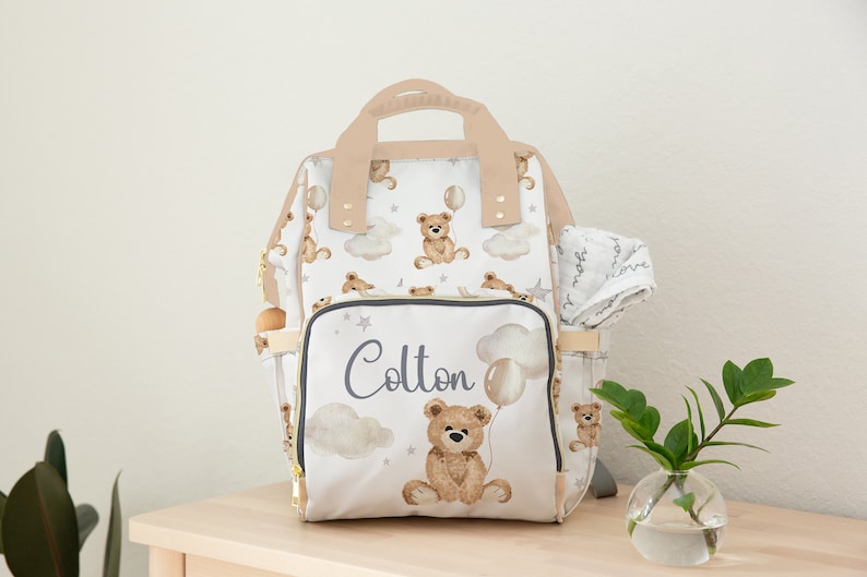 K&ouml;nnte beinhalten: Eine wei&szlig;e Wickeltasche mit einem braunen Teddyb&auml;r- und Ballon-Druck. Die Tasche hat eine Rei&szlig;verschlusstasche mit dem Namen "Colton" darauf. Die Tasche steht auf einem Holztisch mit einer kleinen Pflanze in einer Glasvase.
